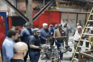 Group of adults in a workshop gathered around a metal piping assembly on a stand, watching a valve demonstration. 