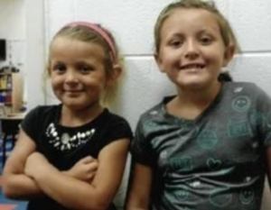 Two young girls stand next to each other, smiling at the camera; one wears a black shirt and pink headband, the other a gray patterned shirt.