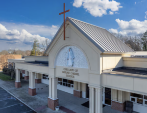 Exterior view of Our Lady of Mount Carmel School church building with a tall cross on the roof and a statue image above the entrance, under a blue sky.