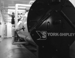 York-Shipley industrial boiler in a mechanical room with an array of pipes and valves surrounding it.