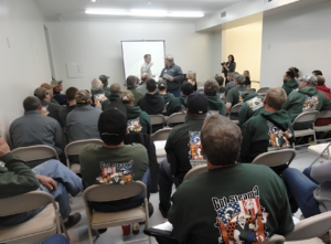 Crowd of men in green hoodies facing a presenter at a projection screen in a bright classroom setup. 