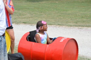 Young girl with pink bow and braided hair sits in a red kayak outdoors, with an adult standing nearby wearing a striped shirt/dress combo.