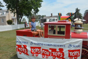Young boy sits on a bright red carnival ride outdoors, smiling, with a large Toys for Tots banner in the foreground and a house and trees in the background.