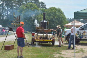Man in red shirt and orange cap watches a steam-powered engine on a trailer at an outdoor event.