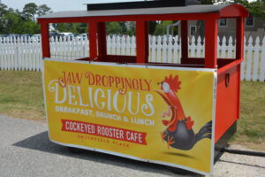 Bright red and yellow food trailer with a large sign reading 'Jaw Droppingly Delicious' and a cartoon rooster graphic outside a parking lot fence.