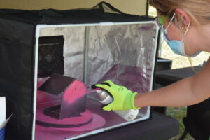 Person wearing a mask and safety goggles spray-paints a pink object inside a small framed tent with reflective foil walls.