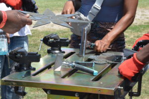People assemble a star-shaped metal sculpture at an outdoor workshop, using clamps and a vise.