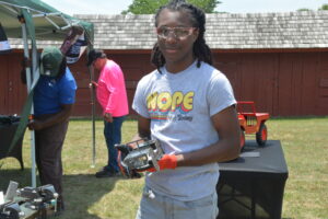 Young man wearing safety goggles and a gray 'NOPE Not Today' T‑shirt, holding a power tool at an outdoor demo.