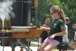 Girl in sunglasses operates a vintage steam-powered machine with brass pipes outdoors.