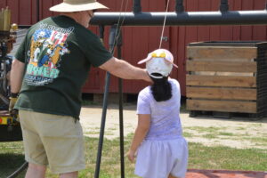 Adult man in a wide-brim hat helps a young girl in a light purple dress outdoors near a large black pipe and wooden crates.