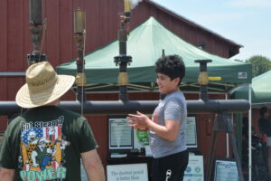 Two men stand near large pipes and a green tent at an outdoor science exhibit, with one wearing a straw hat and a graphic t-shirt and the other in a gray shirt smiling nearby.