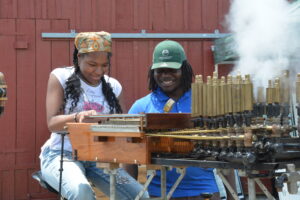Two people play a wooden street organ with brass pipes outdoors, smiling together against a red wooden fence.