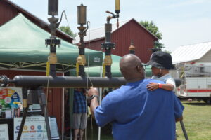 Two men in blue shirts hug beside large industrial pipes at an outdoor event, with a red barn and green canopy in the background.