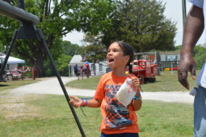 Happy boy in an orange shirt at an outdoor fair, holding a gift bag, with tents and a red fire truck in the background.