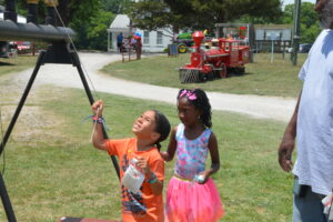 Two young girls at an outdoor event, one in an orange shirt reaching upward and the other in a pink tutu, with a red toy train and fairground backdrop behind them.
