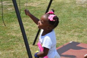 Young girl with pink hair bow stands at a playground, gripping a rope on a swing set outdoors.