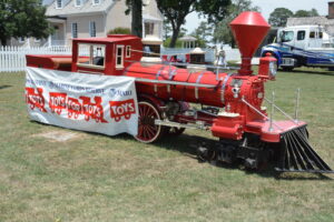 Red vintage steam locomotive on display with a Toys for Tots banner in front, outdoors on grass with a white picket fence and buildings in the background.