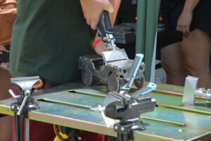 Close-up of a metal model or jig being assembled on a workbench, with a hand gripping a tool and clamps nearby.