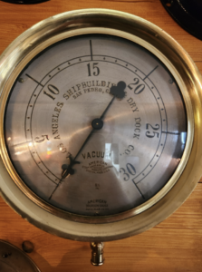 Vintage round vacuum gauge with metal face mounted on a wooden surface; needle points near 15, center labeled VACUUM.