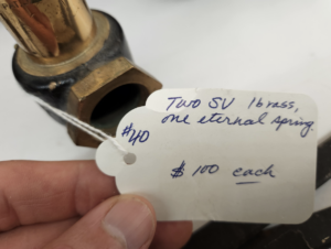 Close-up of a brass valve with a white price tag attached by string, showing $100 each and handwritten notes.