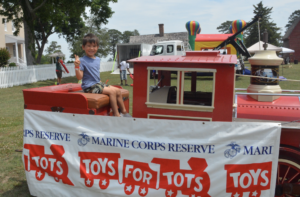 Smiling boy sitting on a red toy train car at a Toys for Tots booth, with banners in the foreground.