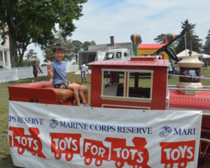 Smiling boy sits on a red toy fire truck at a fair, banner reads 'Toys for Tots' and 'Marine Corps Reserve' in the foreground.