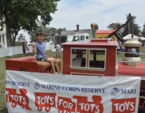 Girl sitting on a red toy train at a Toys for Tots event outdoors, giving a peace sign.