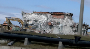 Collapsed concrete building with exposed rebar and debris, an excavator nearby, and a large pipeline in the foreground.
