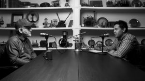 Two men sit across from each other at a long wooden table, speaking into retro microphones in a studio filled with vintage gear on shelves behind them.