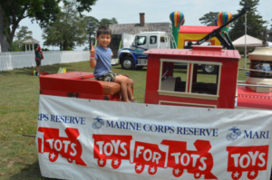 Young boy sits on a red toy train at an outdoor Toys for Tots event, with a banner in the foreground.