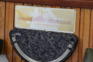 Plaque reading 'U.S. Providence (CL-82), Cleveland-class light cruiser' mounted on a wooden wall above a dusty circular navigational gauge.