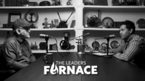 Two men interview each other in a podcast studio with microphones, shelves of trophies behind them; overlay text reads 'The Leaders'.