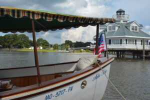 White wooden boat moored at the dock with a striped canopy, an American flag at the stern, and a large white harbor house on pilings in the background.