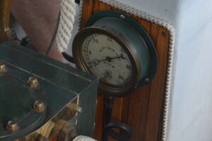 Round pressure gauge mounted on a wooden panel, with rope nearby, showing a scale up to 30 on a vintage boat engine room setup.
