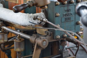 Close-up of a vintage engine component with a braided hose, metal levers, and a teal label reading Tiny Power 2010.