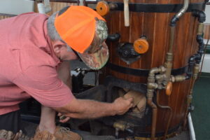 Man wearing an orange cap loads wood into a wood-fired boiler/stove with visible copper piping around it.