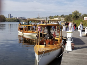 Two vintage wooden boats docked at a riverside pier with a crowd of people mingling along the gangway.