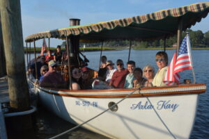 Group of people seated on a striped-canopied boat with an American flag, docked by calm water during a sunny day.