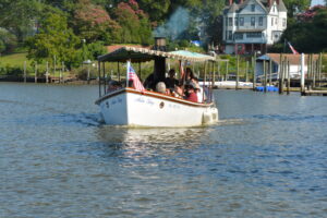 White passenger boat named Arla Kay cruising on a calm river, with an American flag at the bow and passengers under a striped canopy, backed by waterfront houses and trees.