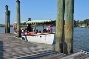 Group of people on a small white boat with a teal canopy docked at a wooden pier; an American flag waves by the side with water and houses in the background.