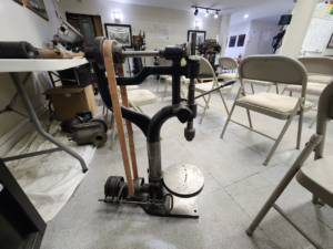 Antique manual exercise/therapy machine with a foot pedal and leather belt on a metal frame in a classroom setting.