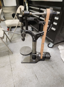 Old metal Canedy drill/press with belt drive and leather strap in a workshop corner.