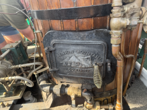 Wood-burning stove with a metal door embossed 'Benson Mountain Company,' surrounded by wood cladding and copper plumbing fittings.