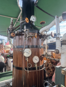 Close-up of a vintage steamship boiler with gauges, valves, and copper pipes on a wooden drum under a green canopy.