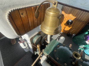 Brass ship bell mounted on a wooden boat wall with coiled rope nearby and water visible beyond.