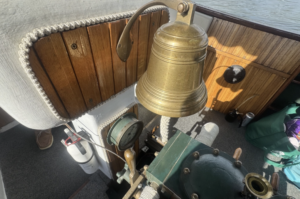 Brass ship's bell hanging above a green helm on a wooden‑paneled boat deck with coiled rope and nautical instruments nearby