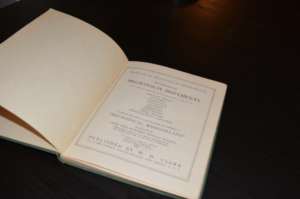 Open vintage book on a dark wooden table, with the title page reading 'Manual of Mechanical Movements'.