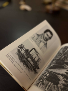 Open book showing a black-and-white illustration of a vintage wagon with a windmill on the left page, and a portrait photo on the right page in the background.