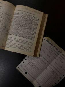 Open old book displaying a dense chart titled 'Properties of Standard Wrought Pipe' with text below; a perforated receipt rests nearby on a dark surface.