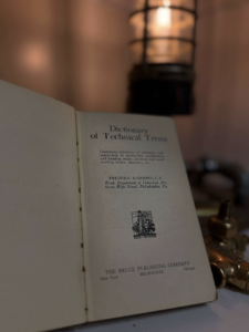 Open page of a vintage Dictionary of Technical Terms with a warm lamp in the background, on a desk.